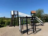 En legeplads med en høj rutsjebane, der ender på et tag i stedet for på jorden, klatrestativer og gynger under en klar, blå himmel.