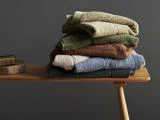 En stak foldede håndklæder i jordfarver - beige, grøn, brun og blå - ligger på en træbænk mod en mørkegrå væg.