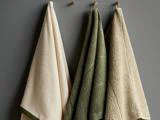 Tre håndklæder - to beige og et grønt - hænger på kroge op ad en grå væg. Hvert håndklæde er struktureret og let draperet.