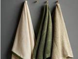 Tre håndklæder - et cremefarvet, et grønt og et beige - hænger på kroge op ad en grå væg.