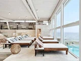 En lys, moderne spa-lounge med liggestole i træ, hvide puder og store vinduer med udsigt over poolen og havet.