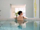 En kvinde med mørkt hår i en knold slapper af i en indendørs pool, læner sig op ad kanten og kigger ind i et lyst, moderne rum med planter.