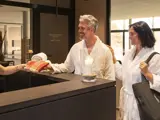 En smilende mand og kvinde i hvide badekåber står ved en spa-reception og modtager håndklæder fra en medarbejder. Et stearinlys i en glasholder står på disken.