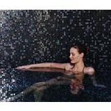 En kvinde i sort badedragt slapper af i en pool med mørke mosaikfliser, hendes arm hviler på poolkanten, og hendes blik er nedadvendt.