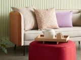 En moderne sofa med forskellige puder står bag en rød ottoman, som rummer en træbakke med tekande og kop. En grøn plante er delvis synlig til venstre.