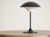 En moderne sort-hvid bordlampe med en kuppelformet skærm sidder på en rund træflade mod en ensfarvet lys baggrund.
