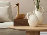 På et moderne sofabord står to hvide keramikvaser med grønne stilke, en andeskulptur i træ og en trææske med en beige sofa i baggrunden.