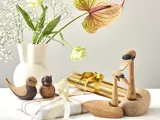 Fuglefigurer af træ og to gaveæsker med guld og hvide bånd står på et bord med en hvid vase med blomster i baggrunden.