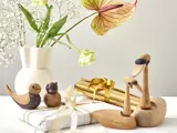 Fugle- og svanefigurer af træ sidder ved siden af hvide og guldindpakkede gaver og en hvid vase med blomster på en lys overflade.