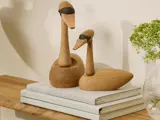 To fuglefigurer af træ med lange halse og sorte øjne sidder på stablede bøger ved siden af en vase med grønne blomster på en træplade.