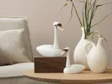 To hvide svanefigurer i keramik på en trækasse ved siden af hvide vaser med tørrede planter på et lyst træbord ved siden af en cremefarvet sofa.