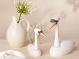 To svanefigurer af træ står på en lys flade ved siden af en hvid keramikkande med grønne blomster og en glasvase med små hvide blomster.