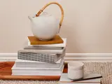 En keramisk tekande med en flettet hank står på stablede bøger ved siden af en keramisk kop og en blyant, alt sammen på en træbakke og et gulvtæppe.