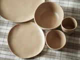 Beige keramiktallerkener, skåle og kopper arrangeret på en ternet dug i neutrale toner.