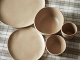 Et sæt beige keramikfade, herunder tallerkener, skåle og kopper, arrangeret på en ternet dug.