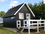 En lille sort træhytte med hvide lister og en veranda foran, omgivet af græs og træer, under en delvis overskyet himmel.