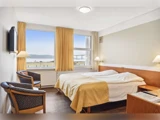 Et hyggeligt hotelværelse med en dobbeltseng, to stole, et lille bord, tv på væggen og et stort vindue med gule gardiner med udsigt over et naturskønt område.