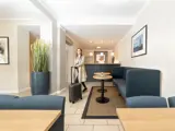 En kvinde i trenchcoat trækker en kuffert gennem en moderne, lys lobby med blå siddepladser, borde og indrammet kunst på væggene.