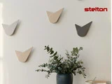 Minimalistiske vægure i neutrale toner er arrangeret på en hvid væg over en grøn vase med eukalyptusgrene; stelton-logoet er i øverste højre hjørne.