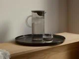 En glaskande med isvand og et glas står på en rund, sort bakke på en lys træplade op ad en almindelig væg.