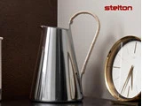 En skinnende kande i rustfrit stål med et buet, indpakket håndtag står ved siden af et rundt ur i guld og hvidt. Ordet stelton står med rødt i øverste højre hjørne.