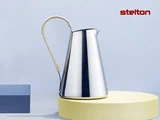 En slank kande i rustfrit stål med et omviklet håndtag står på en gul platform med stelton-logoet i rødt på en lyseblå baggrund.