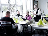 Fire personer sidder omkring et spisebord med foldede grønne servietter, mens en kvinde står og tager imod deres bestilling i en lys, moderne restaurant.