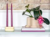 En potteplante med lyserøde blomster står på en stak bøger ved siden af to høje lilla stearinlys i en cremefarvet holder på en hvid hylde med en murstensvæg som baggrund.