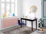Et lyst, moderne arbejdsområde med et sort skrivebord, blå stol, bordlampe, spejl, potteplante og farverig indretning i vindueskarmen over en pink radiator.