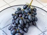 En klase aflange, mørklilla vindruer hviler på en hvid glasplade med et dekorativt mønster.