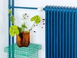 En brun glasvase med grønne og vinrøde blomster står på et grønt trådbord ved siden af en lyseblå radiator på en hvid væg.