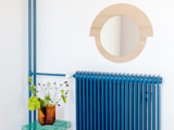 Et rundt træspejl hænger over en blå radiator. På en grøn trådhylde står en ravfarvet glasvase med grønne blade og små hvide blomster.