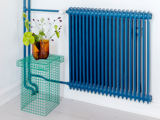 En høj blå radiator med forbundne rør står ved siden af et grønt trådbord med en ravfarvet vase med grønne planter, alt sammen mod en hvid væg og et lyst trægulv.