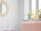 Et lyst rum med et rundt træspejl, en pink radiator, et bord med to blå stearinlys og en vindueskarm med planter og flasker.