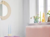 Et lyst rum med et rundt træspejl, en pink radiator, et bord med to blå stearinlys og en vindueskarm med planter og flasker.