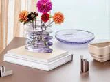 En glasvase med farverige blomster står på en stak bøger ved siden af et lilla glasfad og en beige, bølget skål på et brunt bord.