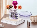 En bølget glasvase med farverige georginer står på stablede bøger ved siden af et lilla glasfad og en beige skål på et lyst træbord ved et vindue.