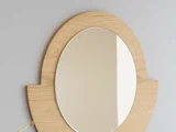 Ovalt spejl med lys træramme monteret på en lysegrå væg, der reflekterer en del af væggen og lyset.