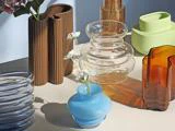 Forskellige farverige glas- og keramikvaser, bl.a. blå, ravfarvede, klare, brune og grønne, udstillet på en neutral overflade med nogle hvide blomster i en blå vase.