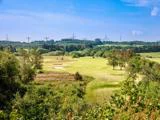 En frodig grøn golfbane med sandbunkers, omgivet af træer og marker; vindmøller og højspændingsledninger kan ses i den fjerne horisont under en blå himmel.