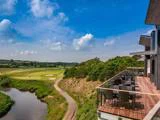 Fra en træterrasse med borde og stole er der udsigt over en golfbane, grønne marker og en snoet sti langs et vandløb under en blå himmel med spredte skyer.