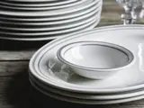 En stak hvide tallerkener og en lille skål med tynde grå kanter står på et træbord ved siden af glas.