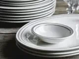 En stak hvide tallerkener med grå kant, en lille skål på toppen og klare glasvarer i baggrunden, placeret på en træoverflade.