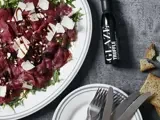 En tallerken med oksecarpaccio med rucola, oliven, høvlet ost, pinjekerner og trøffelglasur står ved siden af bestik, tallerkener, toast og en flaske med trøffelglasur på en grå flade.