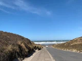 En smal vej og et fortov fører mellem græsklædte klitter mod en sandstrand og havet under en klar blå himmel med en hvid bil parkeret nær kysten.