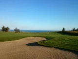 En golfbane med en sandbunker i forgrunden, grønne fairways, træer og en fjern udsigt over havet under en klar blå himmel.