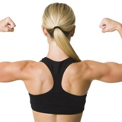 En kvinde med lyst hår i hestehale og iført sort sports-bh spænder sine armmuskler med ryggen til kameraet mod en hvid baggrund.