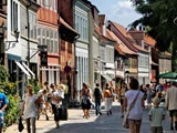 Folk går langs en brostensbelagt gade med farverige, gammeldags bygninger og butikker på en solskinsdag.