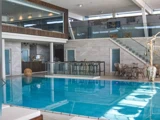 Indendørs poolområde med moderne indretning, trappe med glasgelænder, opholdsområde med borde og stole og stor dekorativ vase ved siden af poolen.