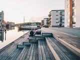 Folk sidder på trætrapper ved en havnefront med oplagte både og moderne bygninger ved solnedgang, hvilket skaber en rolig og afslappet atmosfære.